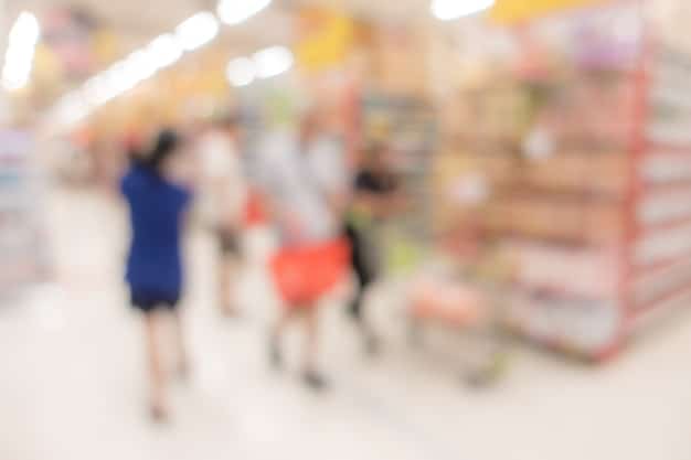 A supermarket aisle scene with blurred shoppers in the background. In the foreground, a hand reaches out to pick up a brightly colored snack box. The focus is on the updated food label on the box, which clearly highlights the added sugar and artificial ingredient content.
